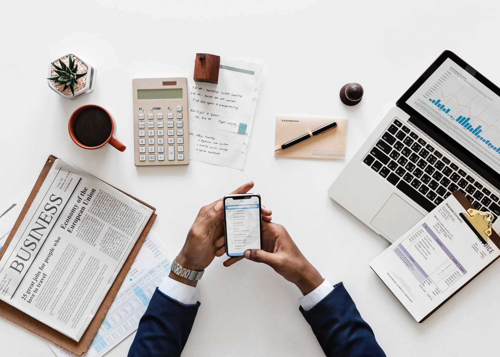 A man with different items, like a laptop, clipboard, pen and paper, calculator, coffee, and a newspaper, using his phone to look up tax information | 2019 Tax Season Changes